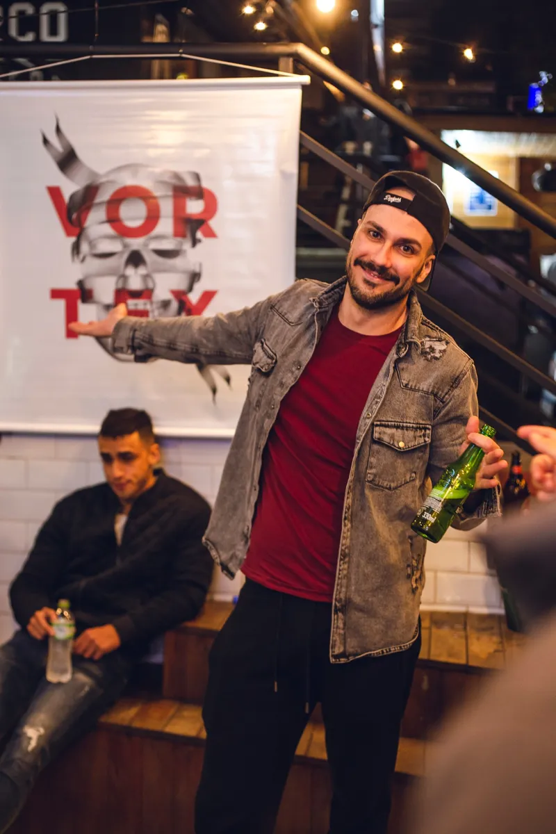 Me presenting the Vortex banner with an open hand, beer in the other, by the staircase.