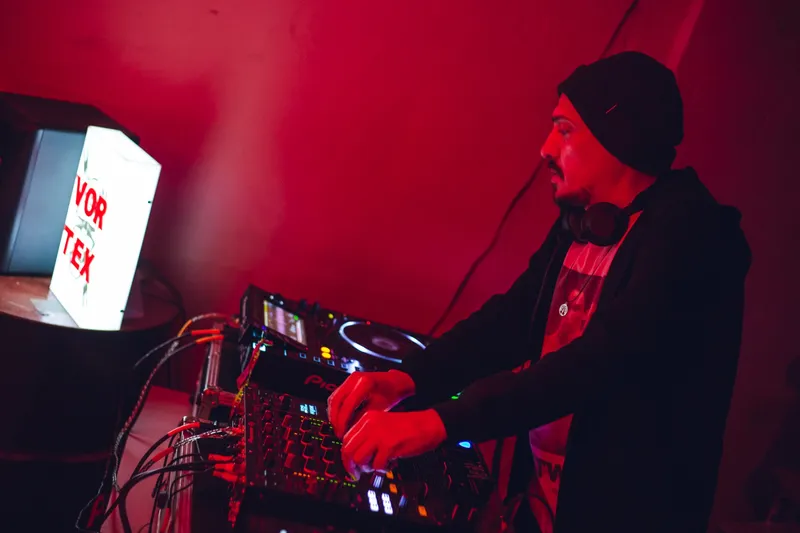 DJ in a beanie mixing under deep red light at a Vortex edition.